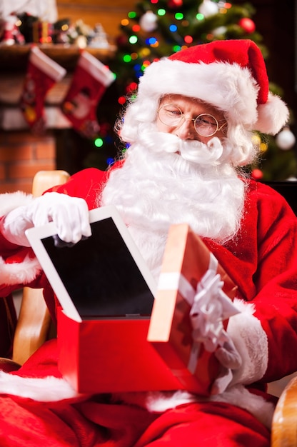 Goed cadeau! Vrolijke kerstman die een digitale tablet in de geschenkdoos stopt en glimlacht terwijl hij op zijn stoel zit met de kerstboom op de achtergrond