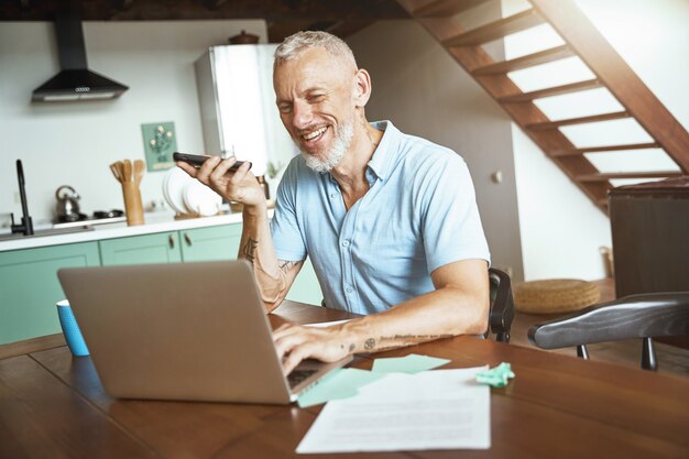 Goed gesprek, gelukkige blanke man van middelbare leeftijd die telefonisch praat terwijl hij aan tafel zit en werkt