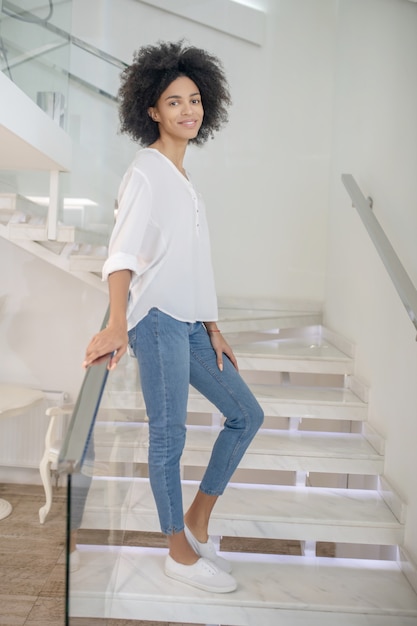 Goed gezind. Gelukkig slanke jonge vrouw in witte blouse leunend op reling op trappen binnenshuis