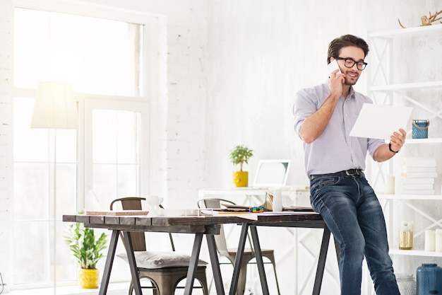 Goed gezind. Knappe waakzame man praten aan de telefoon en met een vel papier