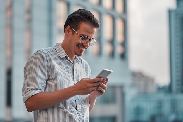 Goed nieuws van collega. Zelfverzekerde jonge man in slimme vrijetijdskleding die een smartphone vasthoudt en ernaar kijkt terwijl hij op straat is.