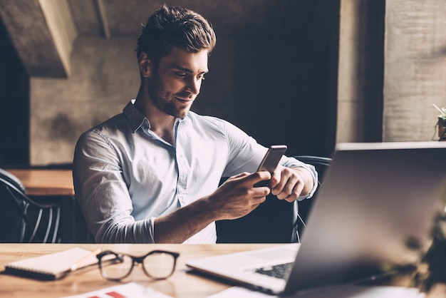 Goed nieuws van collega. zelfverzekerde jonge man in slimme vrijetijdskleding die een smartphone vasthoudt en ernaar kijkt terwijl hij op zijn werkplek op kantoor zit