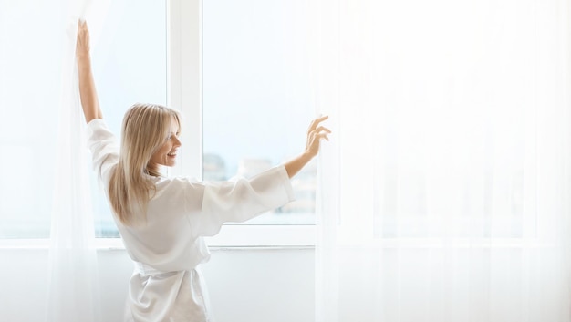 Goed uitgeruste blonde dame die thuis gordijnen opent