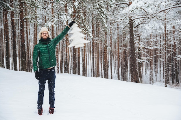 Goed uitziend mannelijk model van middelbare leeftijd draagt een warme bontmuts met oorkleppen en een groene gewatteerde jas met een witte kunstmatige dennenboom staat op de grond bedekt met sneeuw heeft een vrolijke uitdrukking