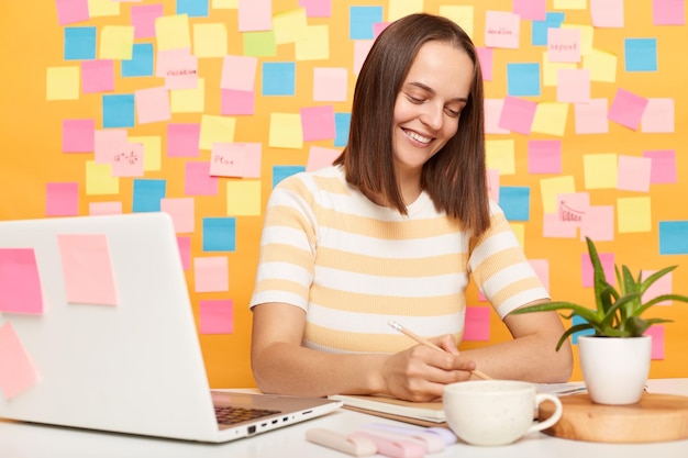 Goed uitziende glimlachende mooie bruinharige zakenvrouw die een gestreept T-shirt draagt, zittend aan tafel op haar werkplek met laptop tegen een gele muur met plaknotities die op papier schrijven terwijl ze haar werk doet