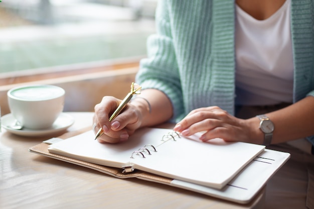 Goed verzorgde vrouwenhand die gouden pen houdt en notities schrijft met gouden pen in notitieboekje terwijl het drinken van blauwe latte naast venster. freelance journalist die thuis werkt. planning toekomstig concept. kopieer ruimte