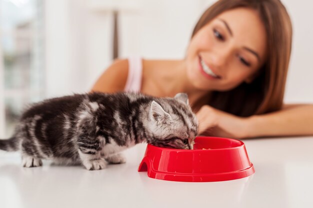 Goed voor haar huisdier zorgen. kleine kitten die voedsel uit de kom eet terwijl een mooie jonge vrouw ernaar kijkt en glimlacht