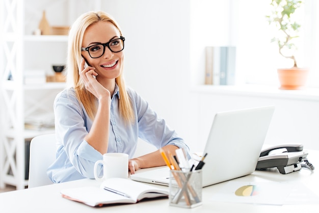 Goed zakelijk gesprek. Vrolijke jonge mooie vrouw in glazen praten op mobiele telefoon en glimlachen terwijl ze op haar werkplek zit