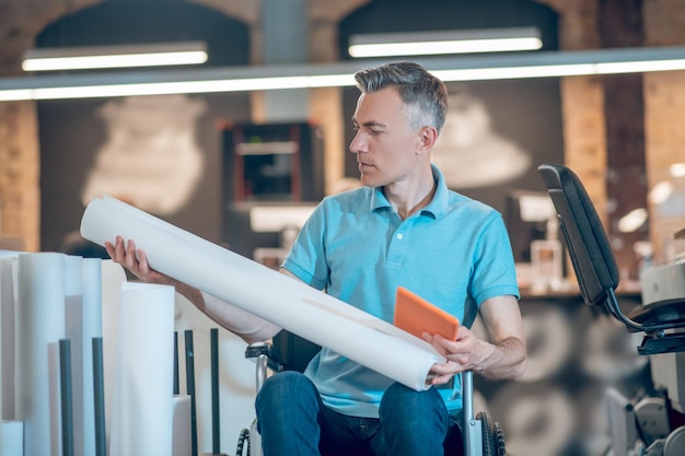 Goede keuze. Volwassen man in blauwe t-shirt op rolstoel met tablet die een rol wit papier in handen onderzoekt en besluit te kiezen in kantoorruimte