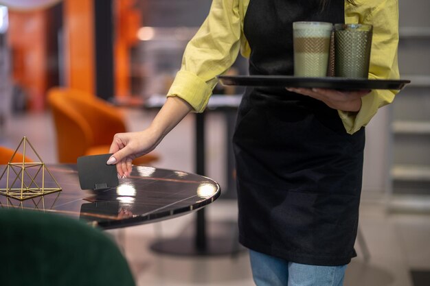 Goede service. Vrouw in schort staande met dienblad met koffie en cappuccino en ontroerende onderzetter op tafel in café geen gezicht