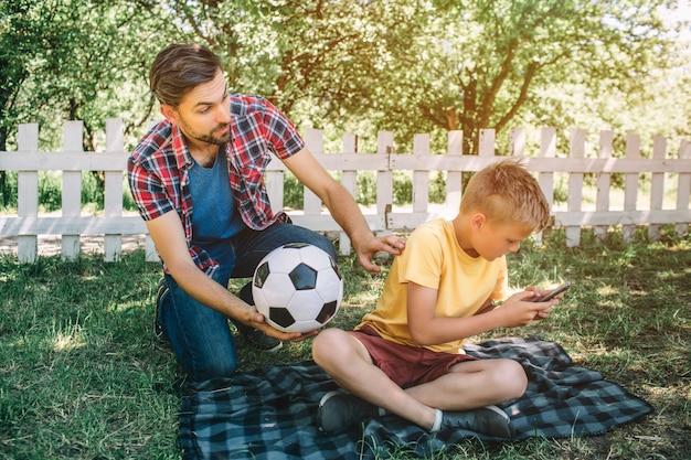 Goede vader kijkt naar zijn zoon en houdt een bal. Hij wil met zijn zoon spelen. Kind speelt op de telefoon. Hij wil geen voetbal spelen.