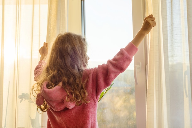 Goedemorgen. Meisje kind in pyjama opent gordijnen