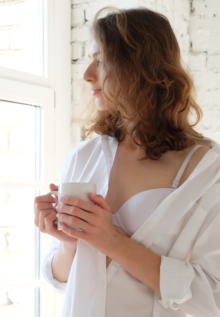 Goedemorgen! mooie vrouw die witte lingerie draagt die 's ochtends koffie drinkt en bij het raam zit.