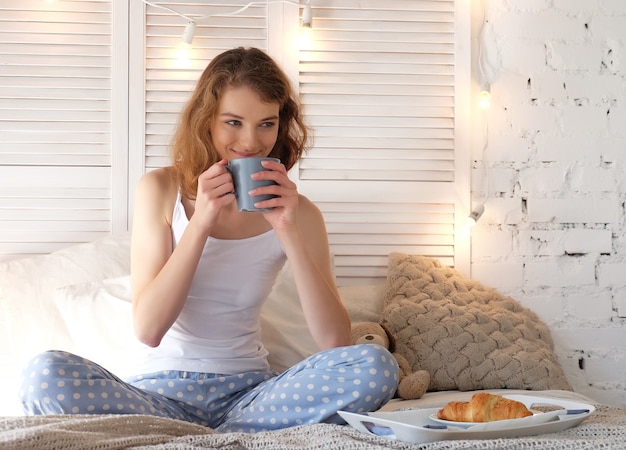 Goedemorgen! Vrolijke vrouw die bovenop bed zit en geniet van haar drankje. Koffietijd. Gezellige slaapkamer.