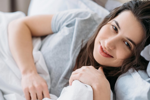 Goedemorgen Weekendrust Thuisontspanning Schoonheidsgezondheid Gelukkige ontspannen luie vrouw die geniet van wakker in bed liggen glimlachend camera kijken in lichte slaapkamer