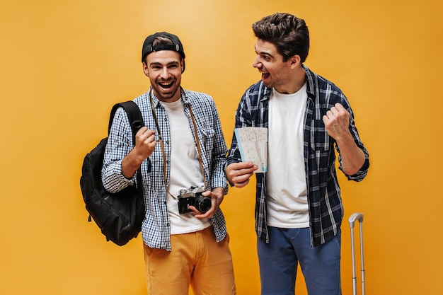 Goedgehumeurde jonge gelukkige reizigers verheugen zich op oranje achtergrond Emotionele gekke mannen in geruite overhemden houden kaartjes en retro camera vast