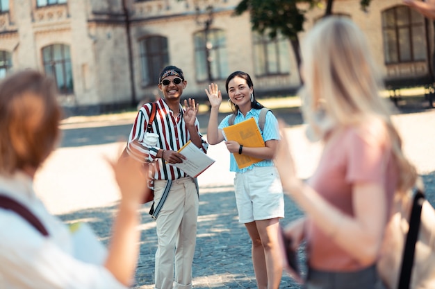 Going to different directions. Two smiling students waving their hands to their classmates standing in the front in university yard.