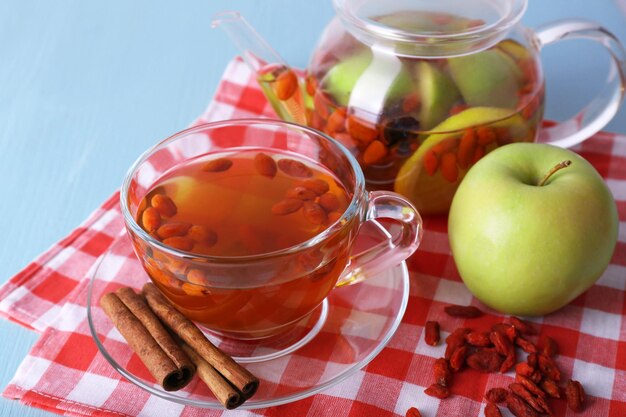 Goji berries drink in glass cup ripe apple and cinnamon fruit drink in glass teapot on red checkered napkin on light blue wooden background