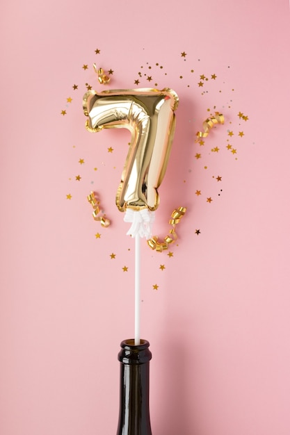 Gold inflatable number 7 on a stick in a champagne bottle surrounded by sequins on a pink background.