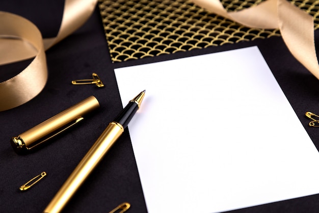 Gold pen, ribbon, paper clips and stationery on a black background with a white sheet of paper with copy space