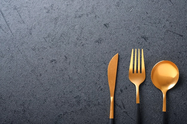 Gold spoon, fork and knife. Set of stylish black and gold cutlery on black background. Fashionable and luxury eating. Top view. Copy space for your text.