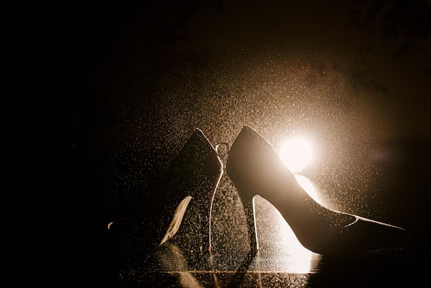小石のあるスタイリッシュな結婚式の靴の間に挟まれた金の結婚指輪暗い背景に水滴のある貴金属の指輪花嫁のスタイリッシュな靴ハイヒールの靴