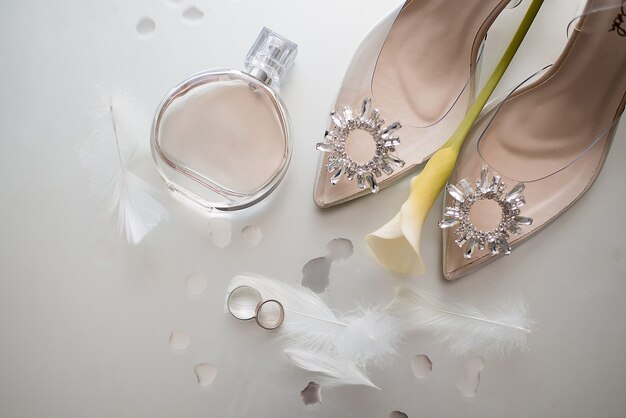 Золотые обручальные кольца с перьями рядом с бежевыми туфлями невесты, украшенными камнями, на которых лежит желтый цветок, и рядом с флаконом духов Chanel