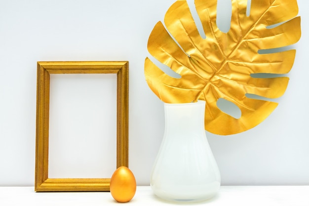 Gold and white interior. Empty photoframe, painted egg and monstera leaf.