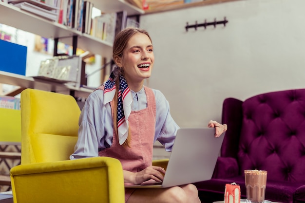 Golden accurate jewelry. Laughing long-haired lady in pink sleeveless dress closing her laptop while having goodies in front