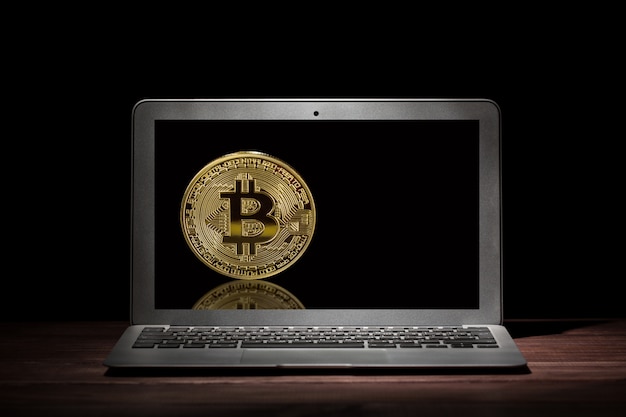 Golden bitcoin on the display of modern laptop in the dark room on wooden table.