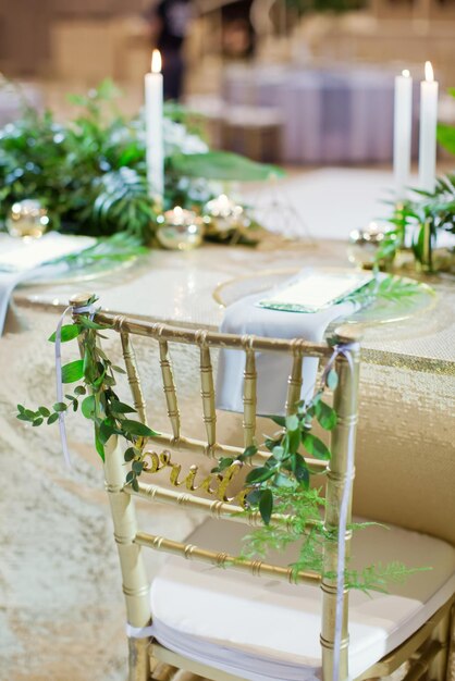 Golden chairs with sign bride and groom with green ruscus twigs Event hall with golden decorations and tropic palm leaves