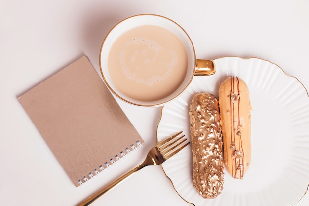 Golden colored eclairs on plate with knife and fork with cup of coffee on light background