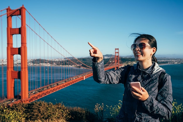 Golden gate Bridge gelukkig wandelen jonge dame toeristische bezienswaardigheden in San Francisco USA. meisje toerist opgewonden wijzend op blauwe lucht met mobiel zoeken in de richting online kaart in Californië VS.