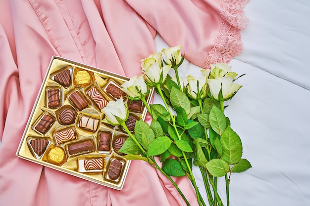 Golden gift box of assorted chocolate on pink silk with white bouquet of rose