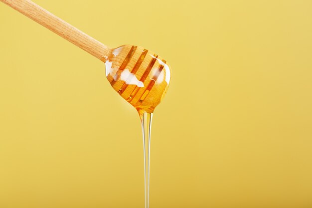 Golden honey trickles from a wooden honey dipper on a yellow background