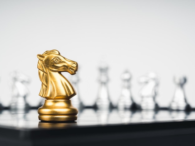 The golden horse knight chess piece stand alone in front of silver chess pieces on chessboard on white background Leadership fighter surviver competition and business strategy concept