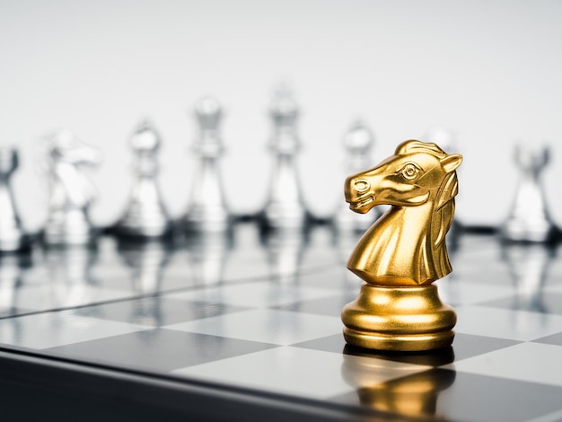 The golden horse knight chess piece stand alone in front of silver chess pieces on chessboard on white background Leadership fighter surviver competition and business strategy concept