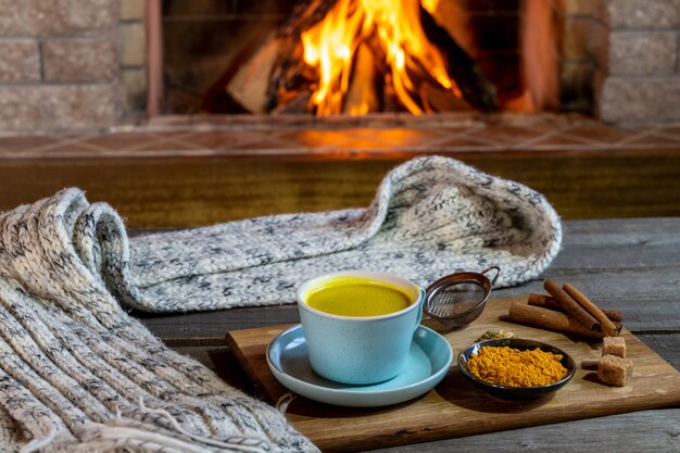 Golden latte milk made with turmeric and spices before cozy fireplace. Healthy coronavirus protection drink.