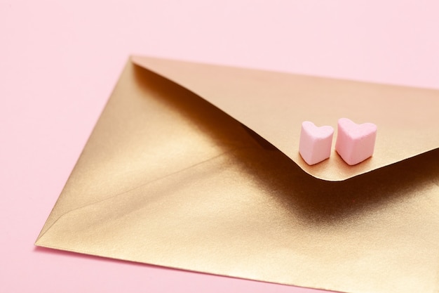 Золотой бумажный конверт с двумя сердечками из зефира на розовом фоне