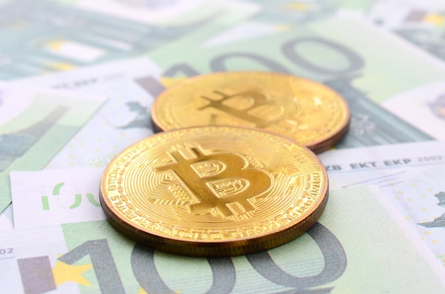 Золотые физические биткойны лежат на множестве зеленых денежных номиналов в 100 евро