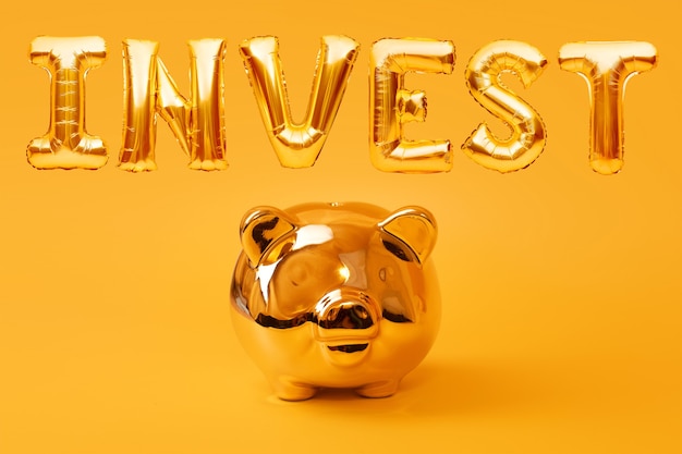 Golden piggy bank on yellow background with golden word INVEST made of inflatable foil balloons. Investment and banking concept. Money saving, moneybox, finance, investments.