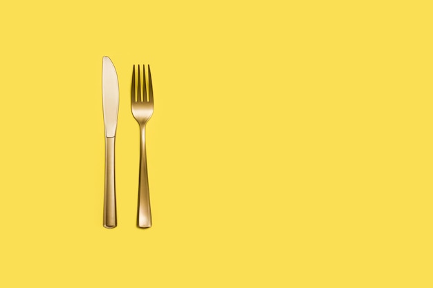 Golden plastic fork and knife on a yellow background with copy space