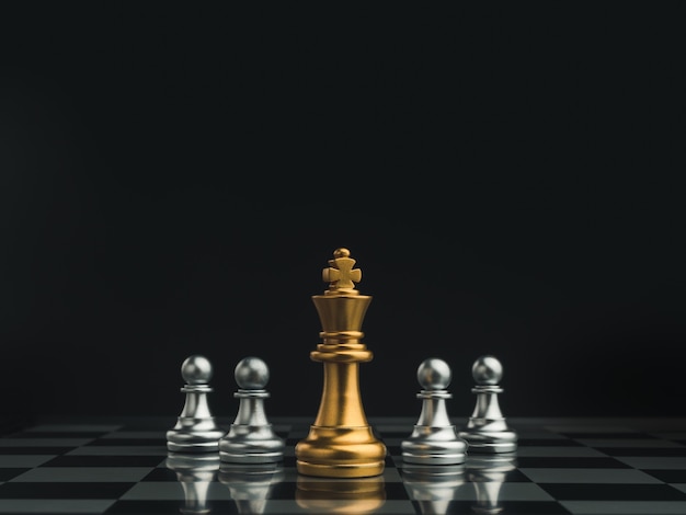 Golden queen chess piece standing in the center of little silver pawn pieces on chessboard on dark background.  Games of chess figure in chess game competition. Leadership, business strategy concept.