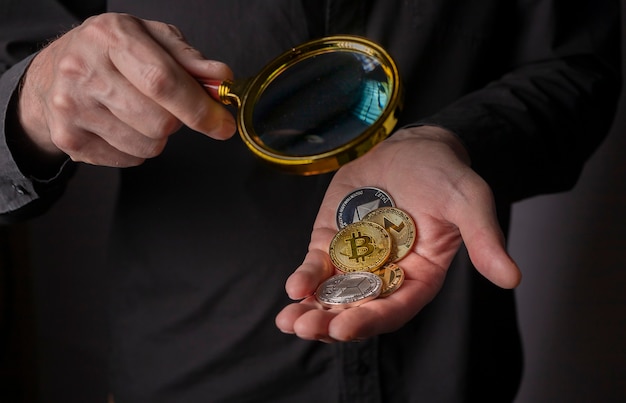 拡大鏡と男性の手のひらの暗号通貨の金と銀のコイン、クローズアップ。ビットコインと他の暗号通貨の山。