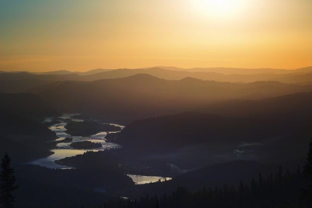 Golden sunset in the mountains: dark silhouettes of the hills, golden light in the haze, clouds in the blue sky, at the bottom of the valley reflection in the water of the river.