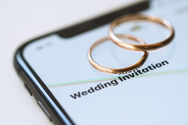 スマートフォンの上にある金色の結婚指輪と画面上のテキスト 結婚式の招待状