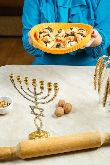 메노라와 함께 테이블에 서 있는 여성의 손에 쟁반에 있는 푸림의 유대인 휴일을 위한 전통적인 고멘타시 쿠키