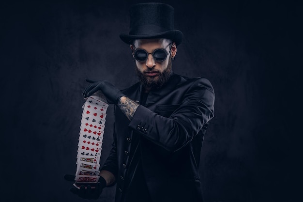 Goochelaar in een zwart pak, zonnebril en hoge hoed, met truc met speelkaarten op een donkere achtergrond.