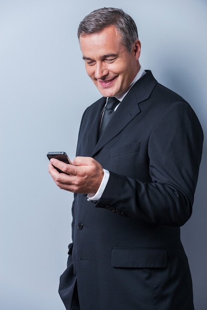 Хорошие деловые новости. Уверенный зрелый мужчина в строгой одежде держит мобильный телефон и смотрит на него с улыбкой, стоя на сером фоне