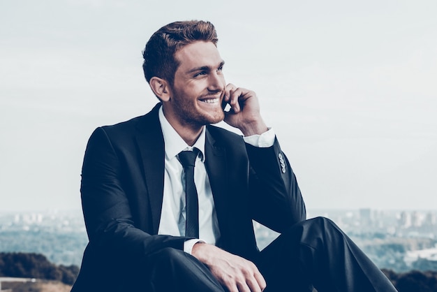 Хороший деловой разговор. Уверенный молодой человек в строгой одежде разговаривает по мобильному телефону и улыбается, сидя на открытом воздухе на фоне городского пейзажа